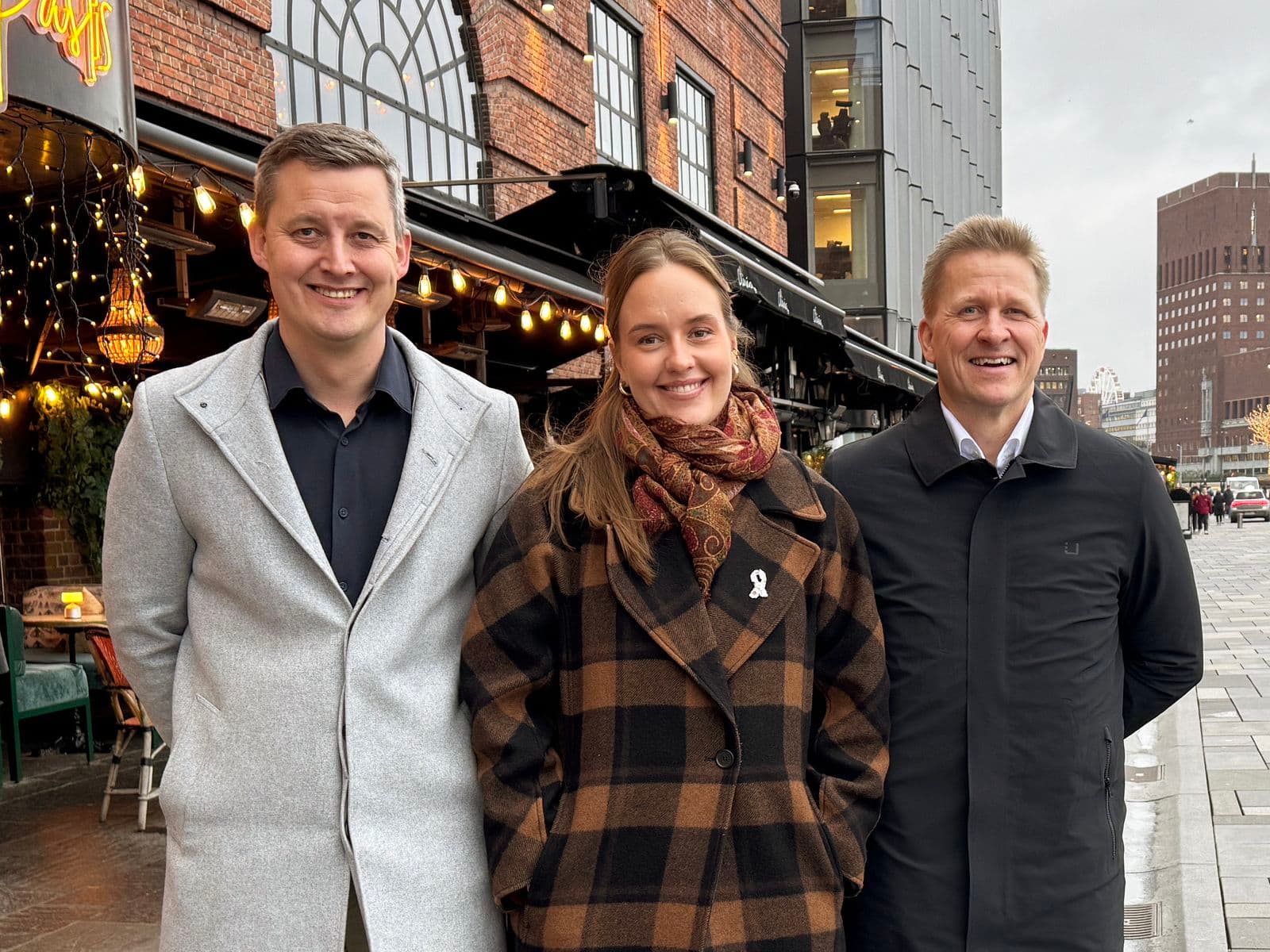 Three people, two men and a woman, smiling and standing on a city street with buildings in the background on an overcast day.