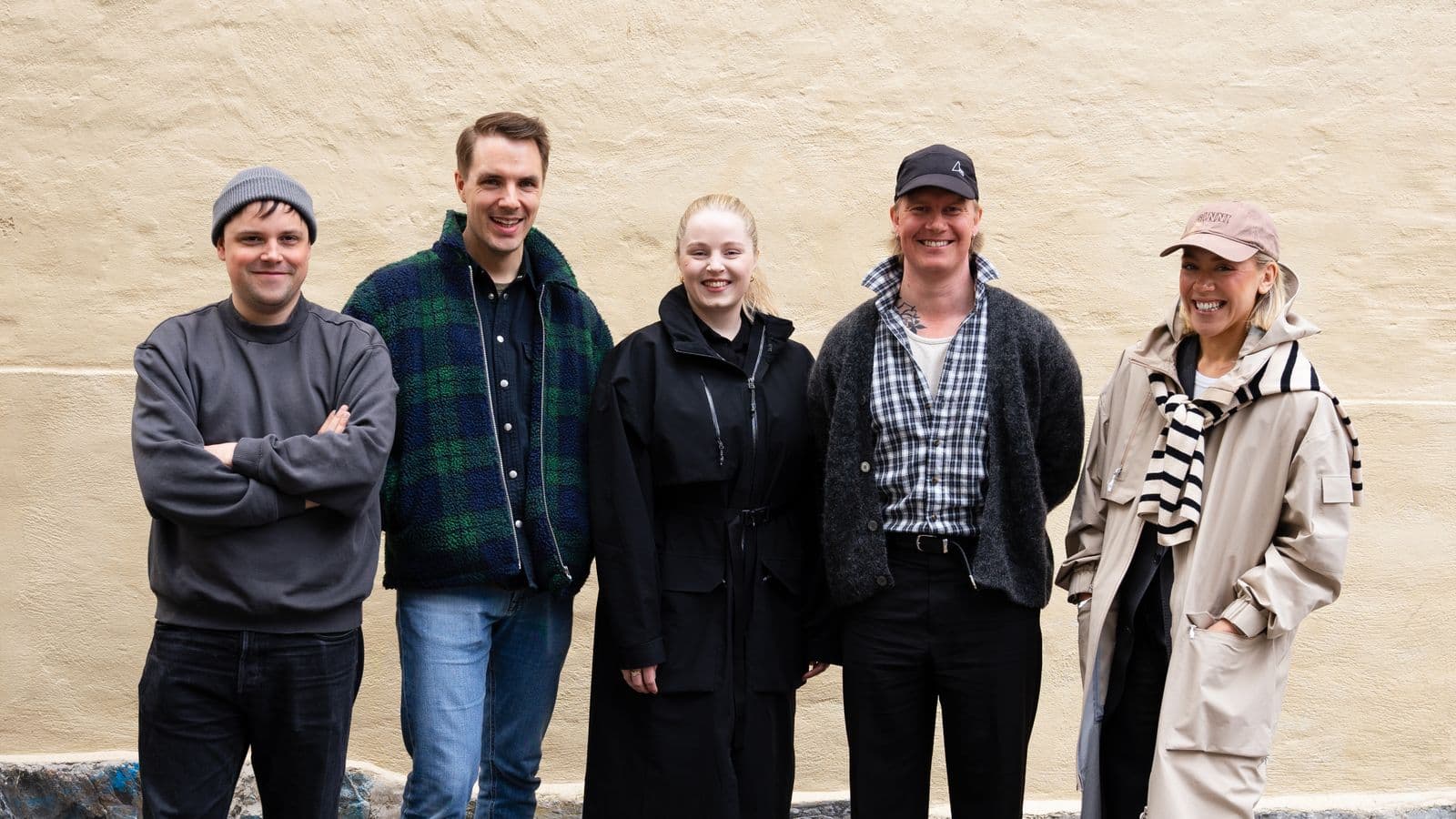 Five people standing in a row, smiling in casual outfits against a beige wall background.